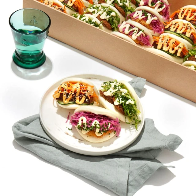 Close-up of bao buns filled with assorted toppings on a plate, alongside a glass of water and a box of more bao buns.