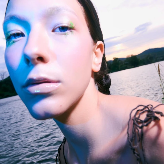 Close-up of fair-skinned young adult woman near a lake at sunset with blue skin lighting, shimmering eye makeup, and textured dress.