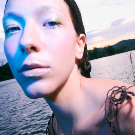 Close-up of fair-skinned young adult woman near a lake at sunset with blue skin lighting, shimmering eye makeup, and textured dress.