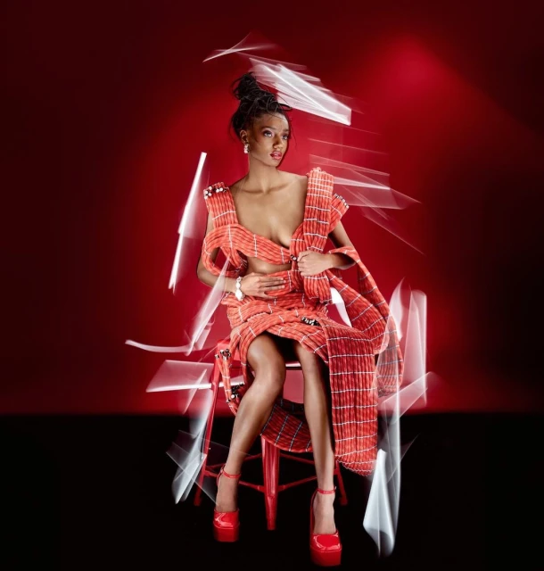 A stylish woman with an updo hairstyle is posing in a red and white plaid dress and red heels against a red background.