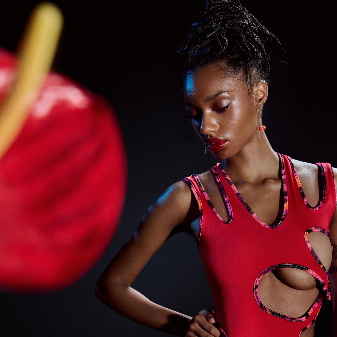 Close-up of a young Black woman posing in a red leotard with cutouts against a dark background, accented by a blurred red flower.