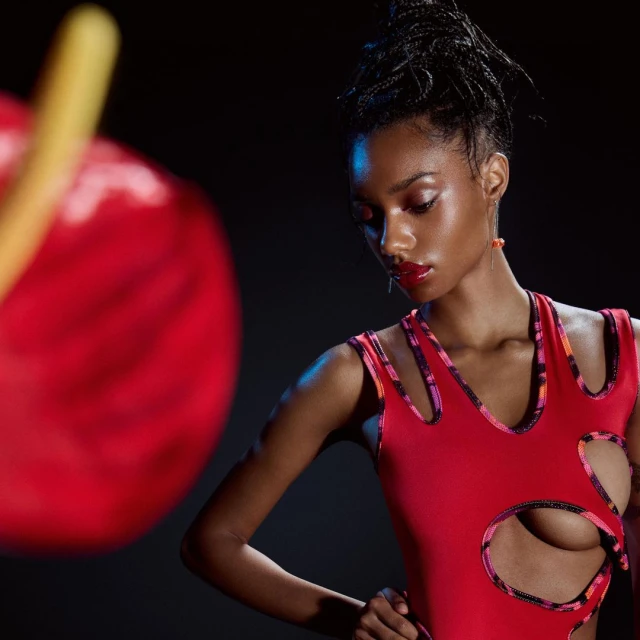 Close-up of a young Black woman posing in a red leotard with cutouts against a dark background, accented by a blurred red flower.