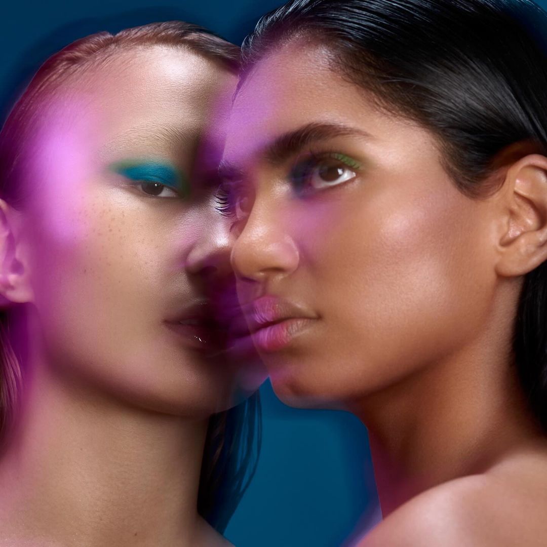 Close-up beauty shot of two diverse adult females with blue and green eyeshadow, diffused lighting, against a dark teal background.