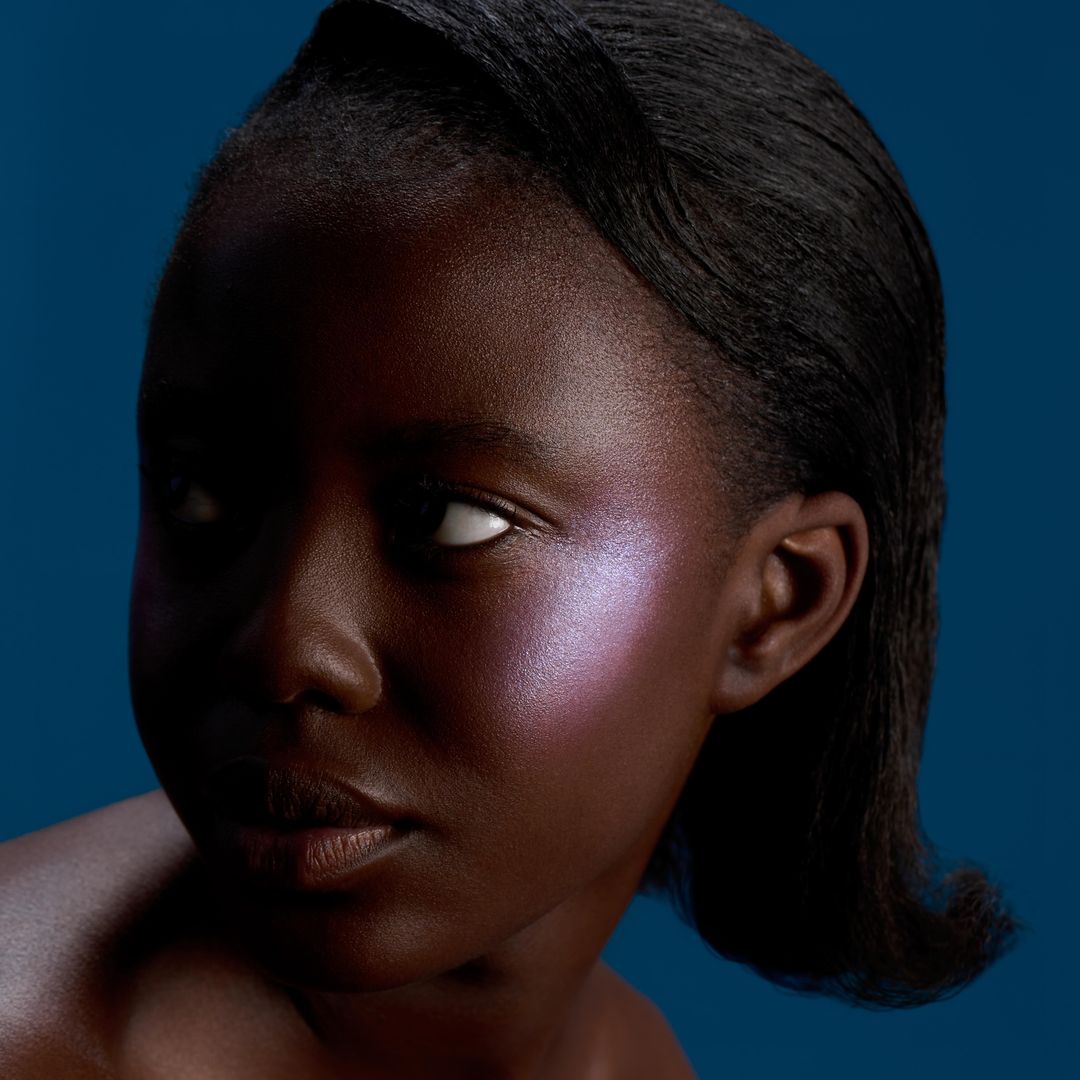 Portrait of a young Black woman with violet makeup on her cheekbones, looking to the side, with dark hair against a blue background.