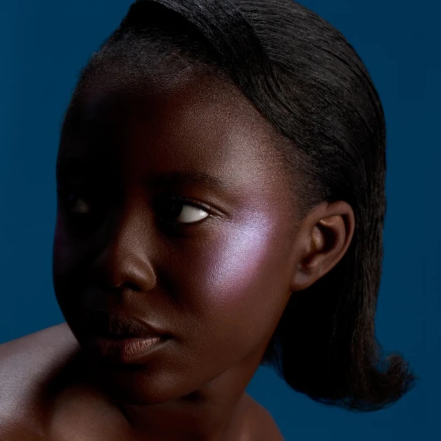 Portrait of a young Black woman with violet makeup on her cheekbones, looking to the side, with dark hair against a blue background.