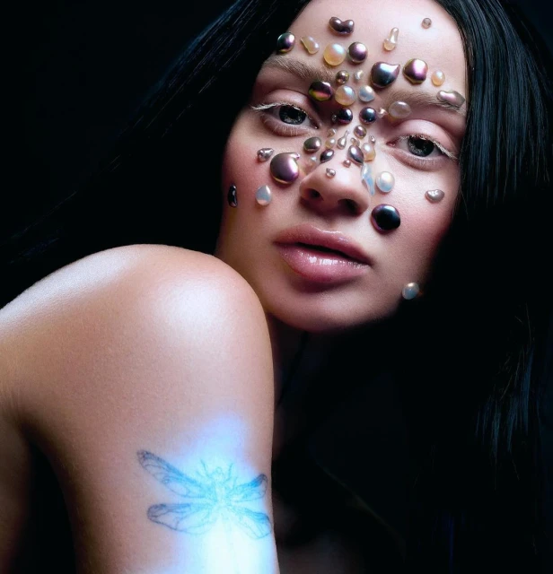 Close-up studio portrait of a woman with iridescent pearls applied to her face and shoulder, dark hair, light skin, dark studio background.