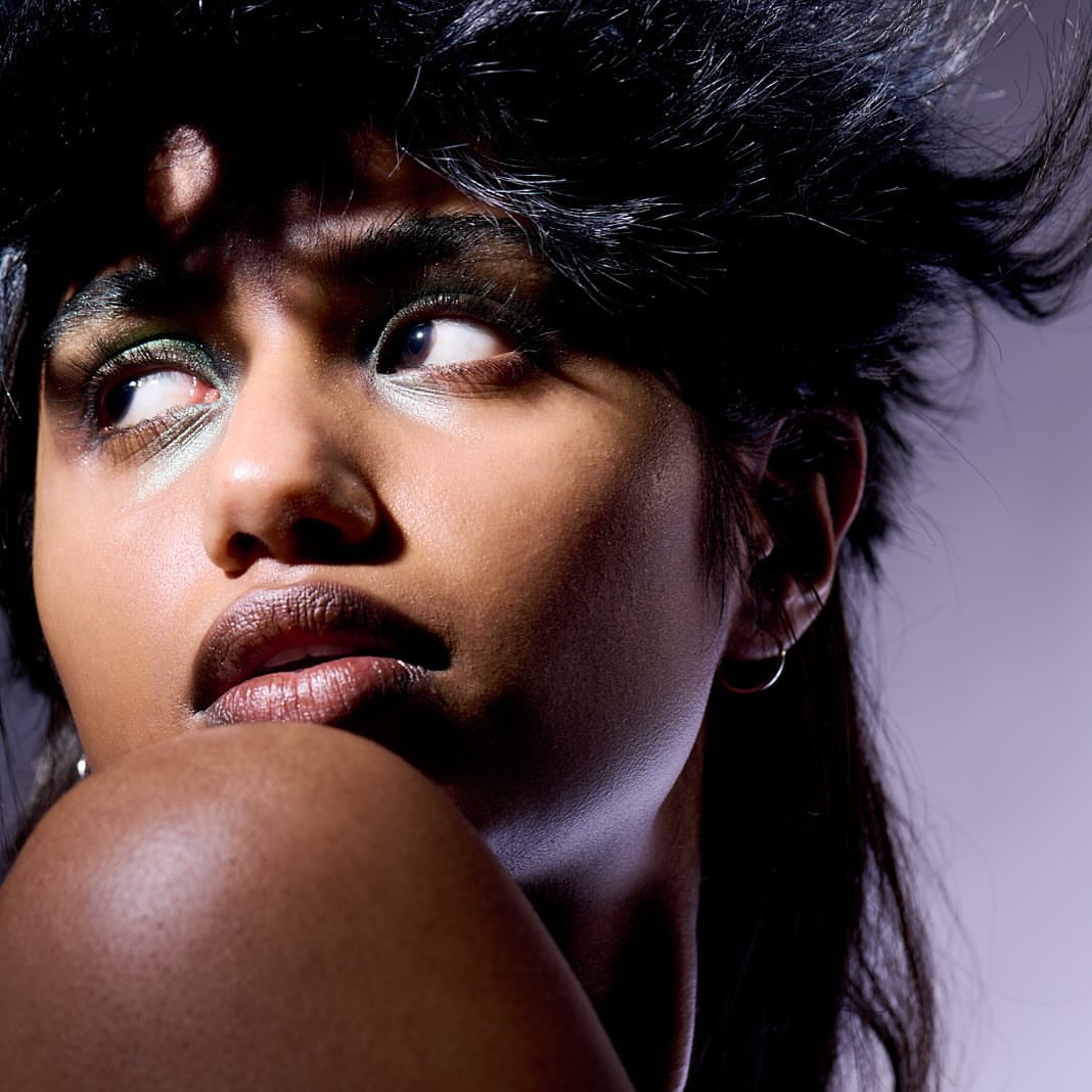 Close-up beauty shot of a woman with dark hair and green eye shadow looking up and away from the camera in a studio setting.