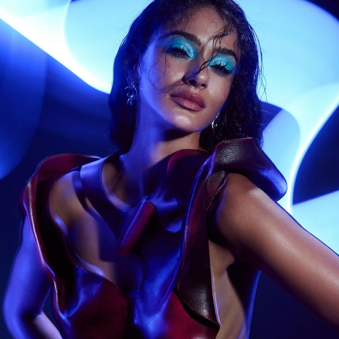 Portrait of a woman with blue eyeshadow, wearing a sculptural dress, posing against a blurred blue light background in a studio.