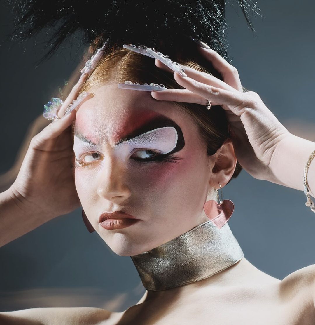 Close-up of a fair-skinned adult female with red hair and creative theatrical makeup, wearing statement earrings and a metallic choker.
