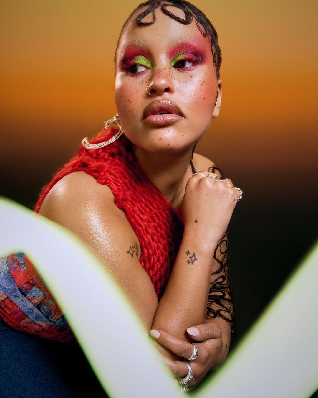 Stylish portrait of a woman with creative hair design, makeup, and tattoos, posing with hands together, wearing a red knit top.