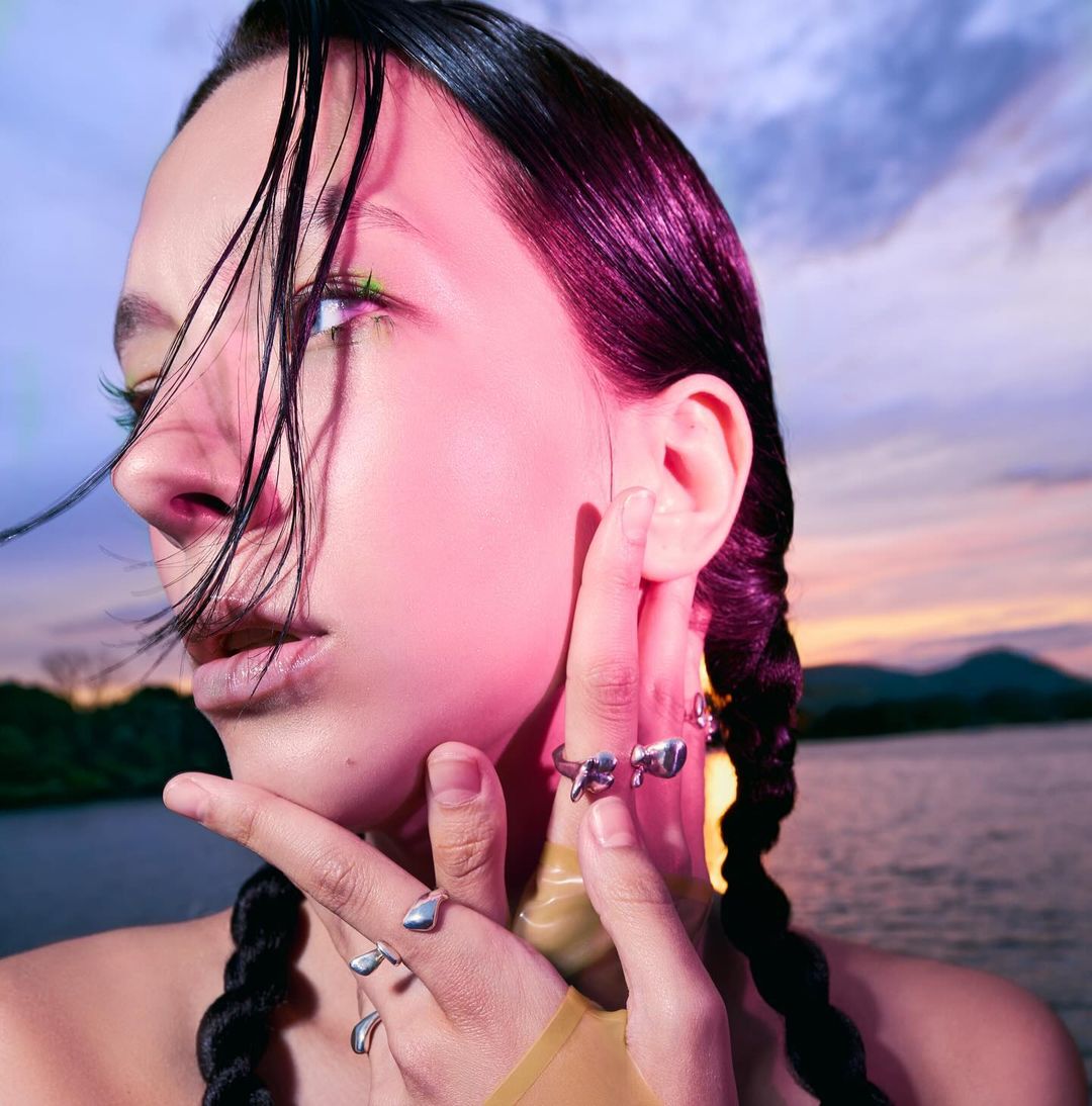 Close-up of a young woman with braided hair, wearing rings and pink eyeshadow, posing in front of a lake at sunset with mountains.