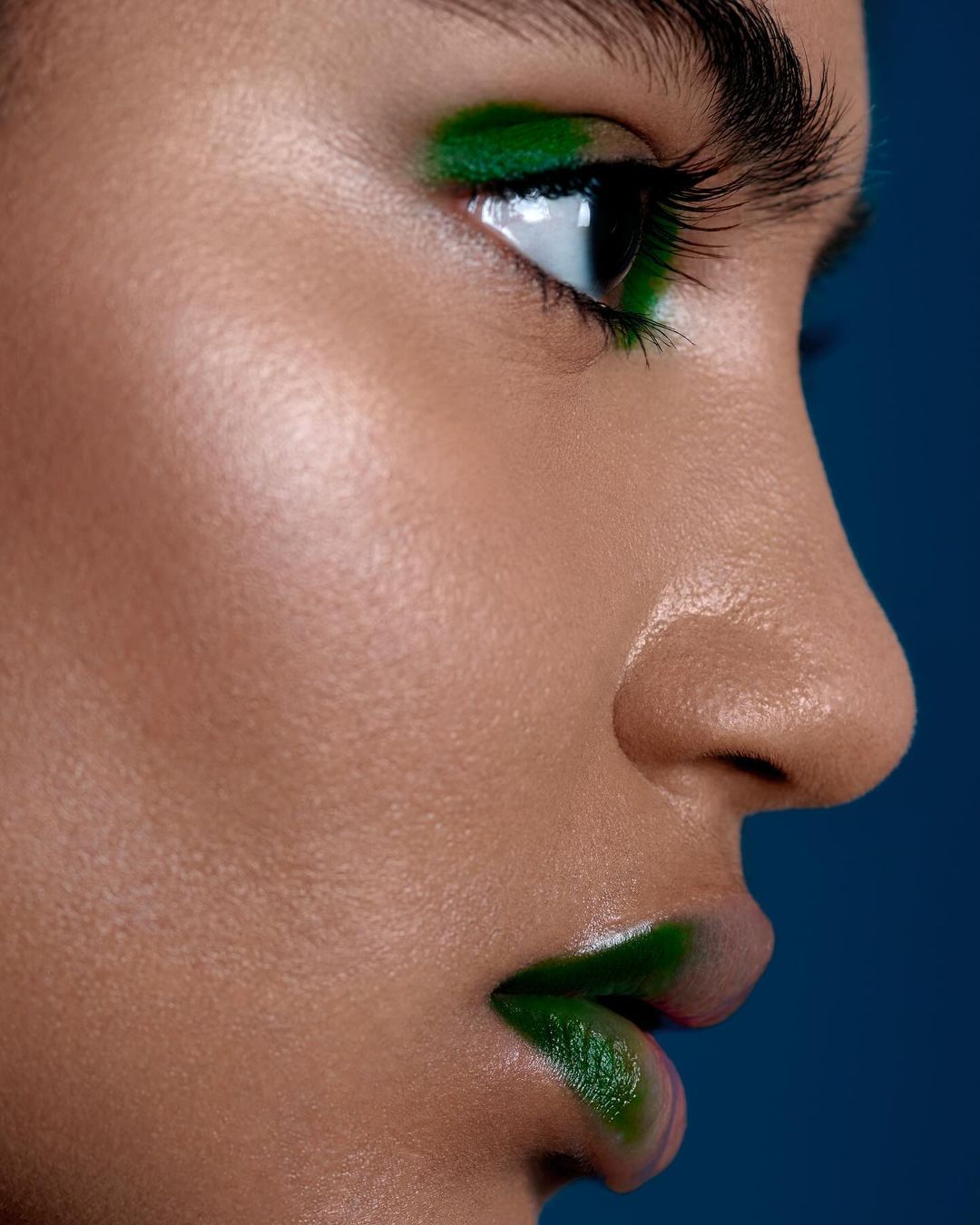 Close-up profile of a young adult female with dark hair and olive skin, wearing green makeup on her lips and eyelids against a blue background.