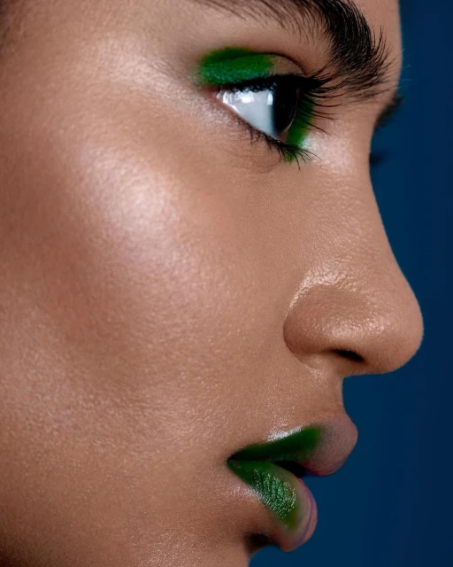 Close-up profile of a young adult female with dark hair and olive skin, wearing green makeup on her lips and eyelids against a blue background.