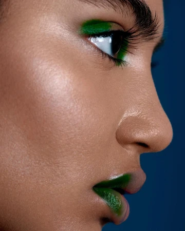 Close-up profile of a young adult female with dark hair and olive skin, wearing green makeup on her lips and eyelids against a blue background.