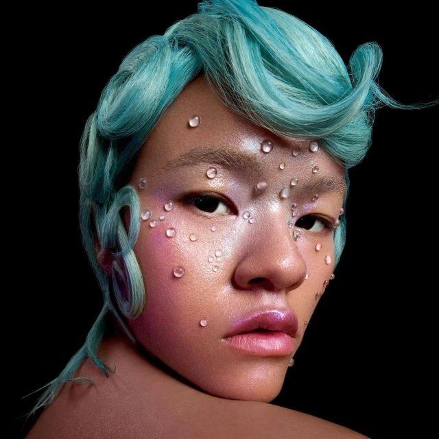Close-up portrait of a young adult with blue stylized hair, iridescent makeup, and water droplets on the face against a dark backdrop.