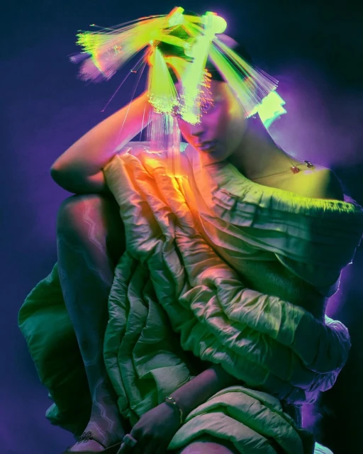 Woman in artistic fashion pose with illuminated fiber optic headpiece, draped in draped light-green fabric, against dark purple backdrop.