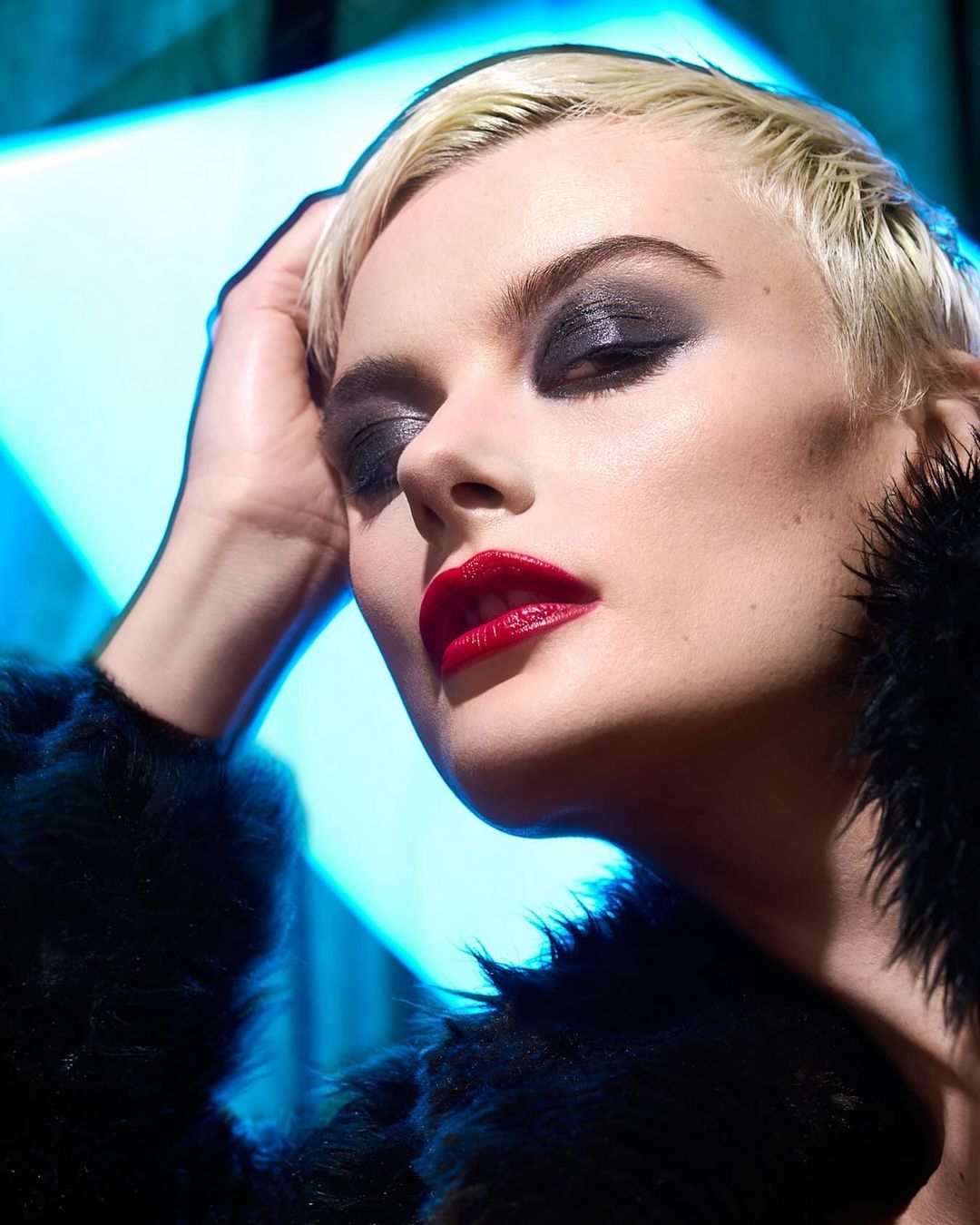Close-up of a woman with blonde hair, dark eyeshadow, and red lipstick, wearing a black fur coat against a bright blue background.