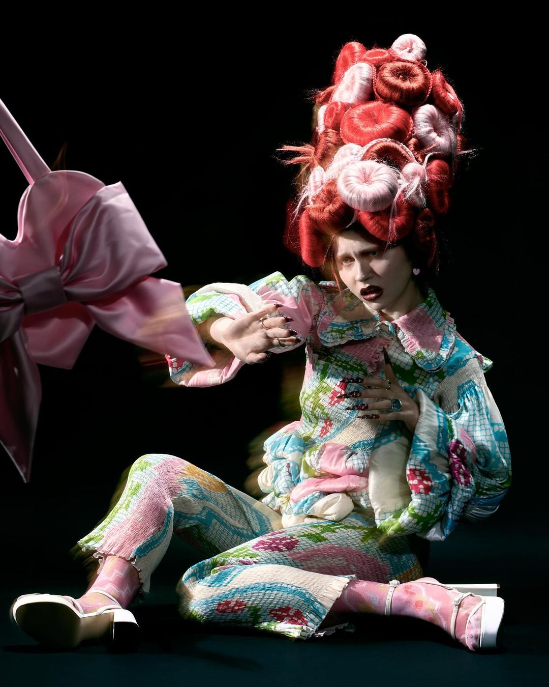 Fashion photo of a posed adult female wearing a floral patterned outfit with a large wig and platform heels, seated in a dark studio.