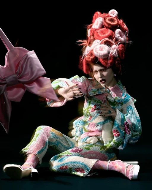 Fashion photo of a posed adult female wearing a floral patterned outfit with a large wig and platform heels, seated in a dark studio.