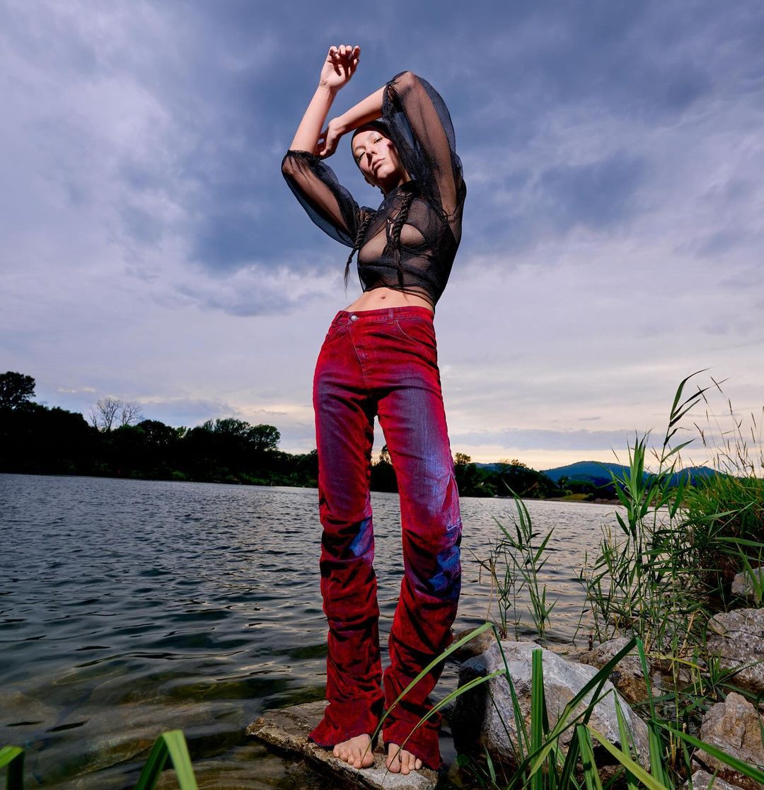 A tall, slender adult female model in a sheer black blouse and patterned red pants poses on rocks near a lake with an overcast sky.