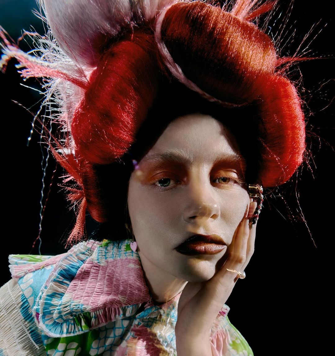 Close-up portrait of a woman with elaborate red and white hairstyle, vibrant makeup, and a colorful patterned top against a dark background.