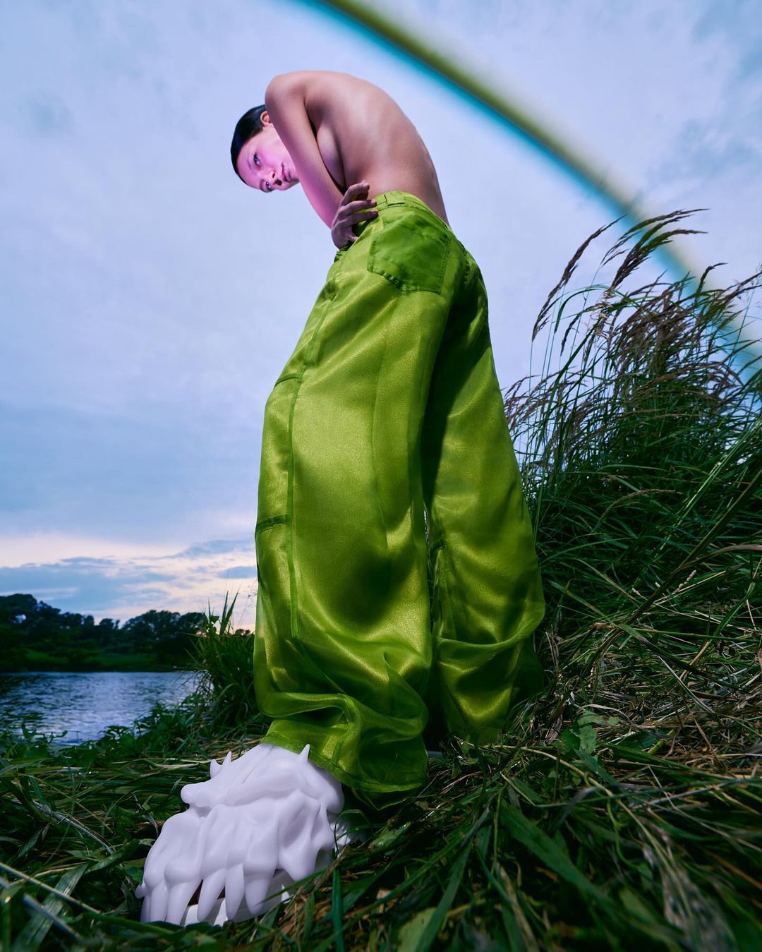 A bare-chested person in green satin pants stands in grass near a river under a blue sky with a rainbow, looking down at the camera.