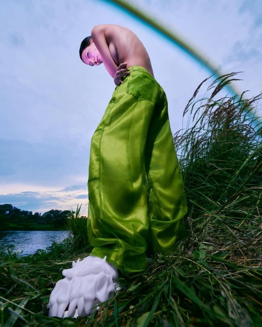 A bare-chested person in green satin pants stands in grass near a river under a blue sky with a rainbow, looking down at the camera.