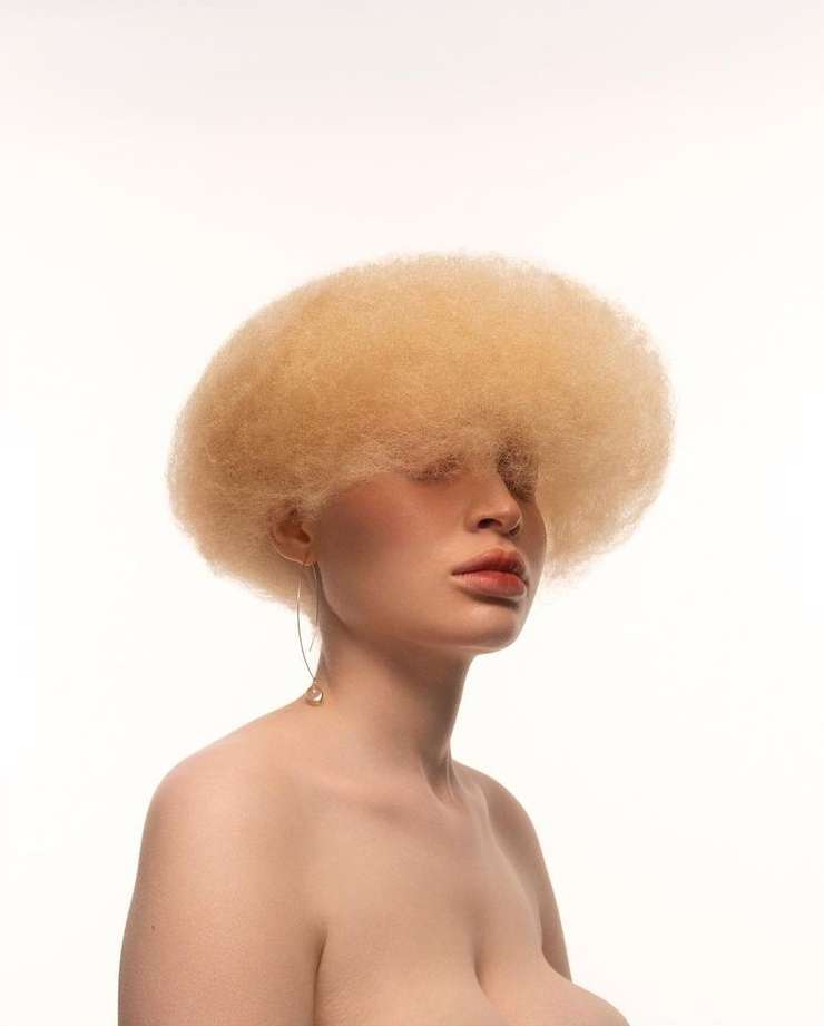Close-up studio portrait of an adult female with albino skin, voluminous afro hairstyle, and minimal makeup on a clean white backdrop.