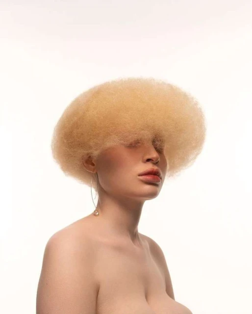 Close-up studio portrait of an adult female with albino skin, voluminous afro hairstyle, and minimal makeup on a clean white backdrop.