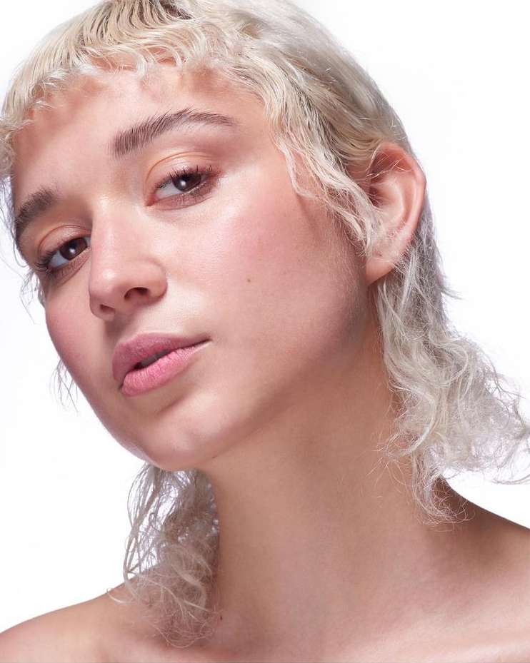 Close-up portrait of a young adult woman with curly blonde hair and pink lipstick against a bright white backdrop in a beauty shot.