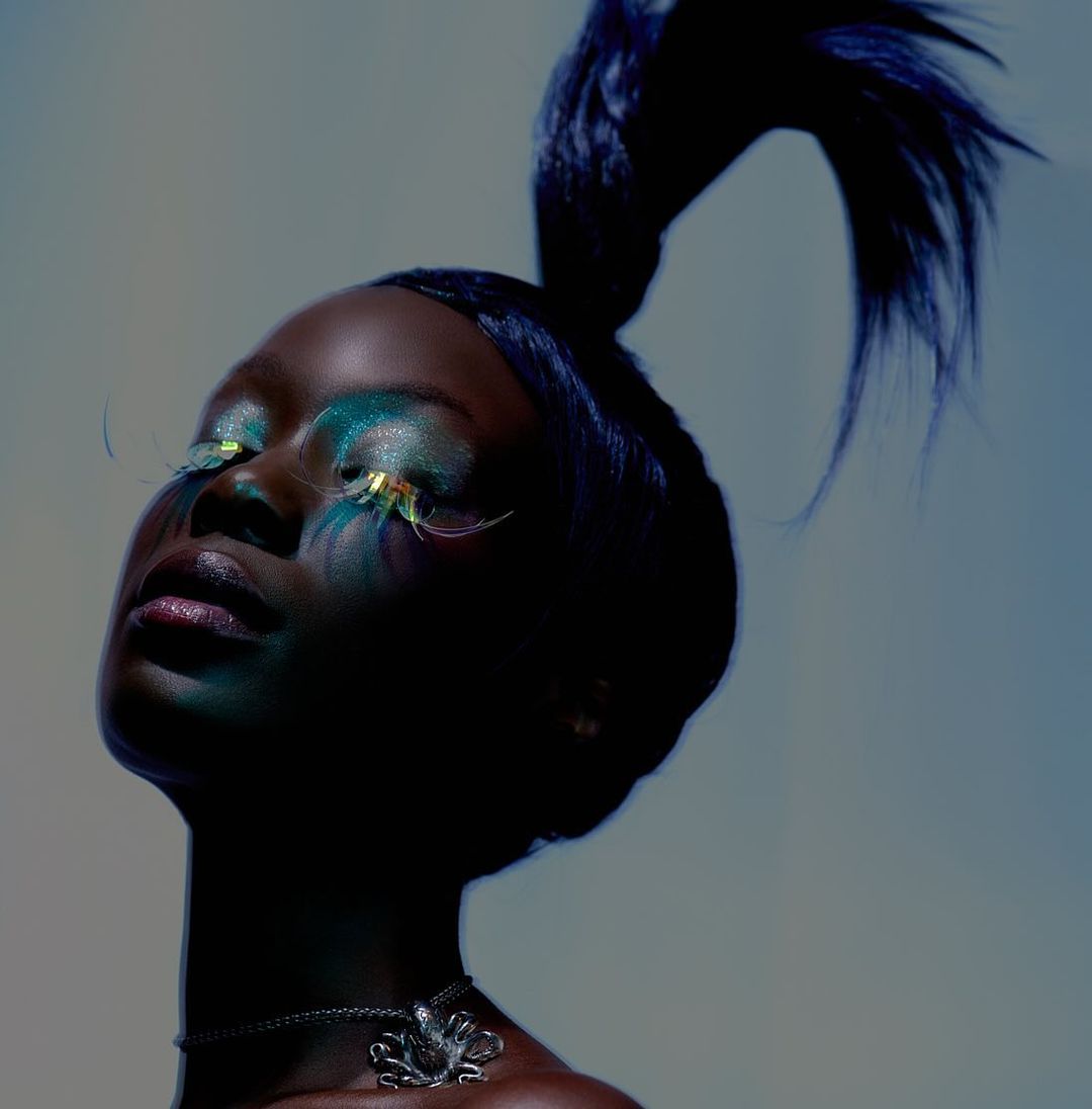 Beauty shot of a Black woman with elaborate makeup, false eyelashes, and an octopus pendant against a gradient blue background in studio.