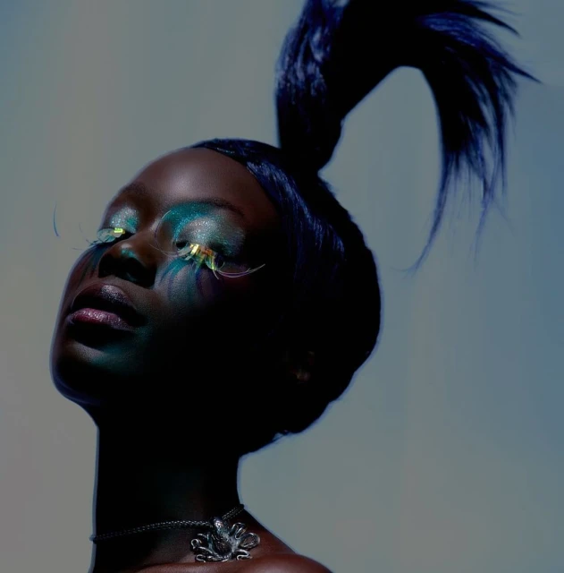 Beauty shot of a Black woman with elaborate makeup, false eyelashes, and an octopus pendant against a gradient blue background in studio.