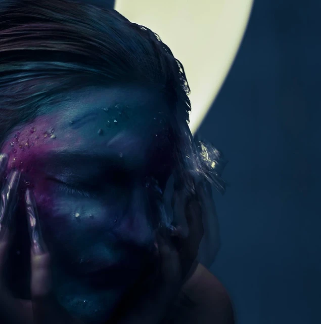 Portrait of a woman with wet hair and face makeup, obscured by plastic wrap against a blue and cream backdrop with soft lighting.