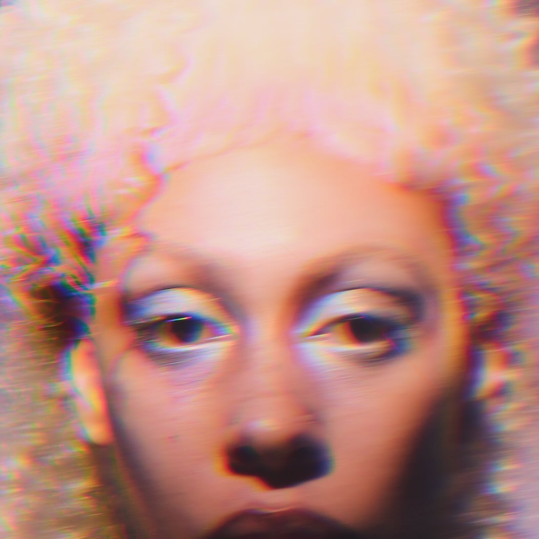 Close-up of an adult female with light blonde afro-style hair and dramatic makeup, digitally distorted with blurred and chromatic aberration effects.
