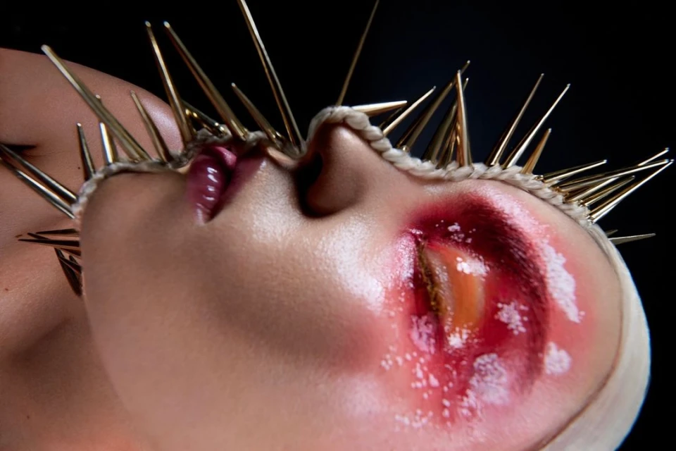 Artistic beauty portrait of a woman with a crown of gold spikes, dramatic eye makeup, and glossy lips against a dark background.
