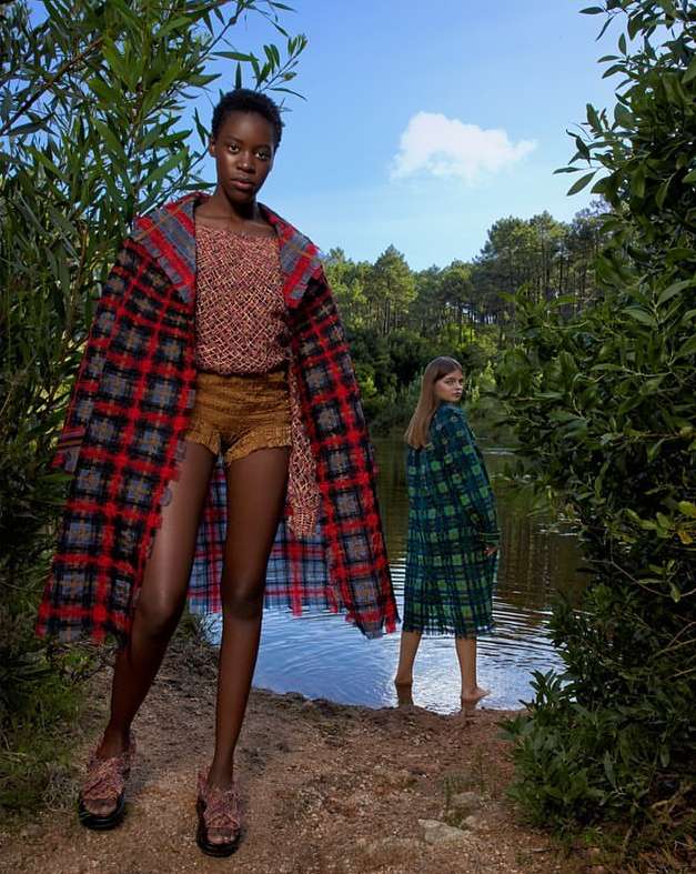 Two fashion models showcase outerwear near a lake; an African woman in a red plaid coat stands while a Caucasian woman poses in green.