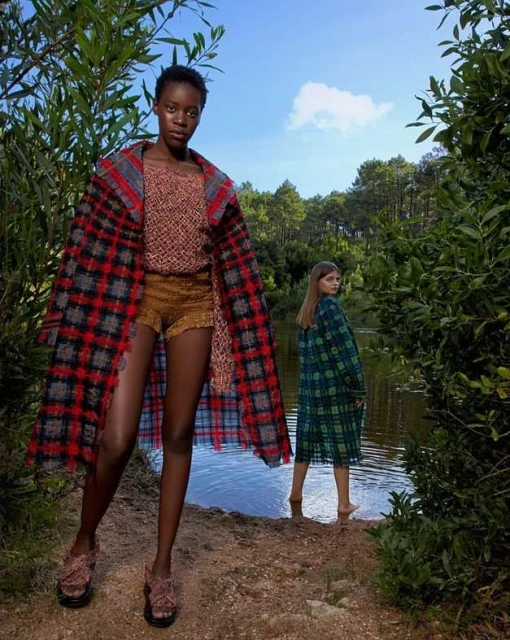 Two fashion models showcase outerwear near a lake; an African woman in a red plaid coat stands while a Caucasian woman poses in green.