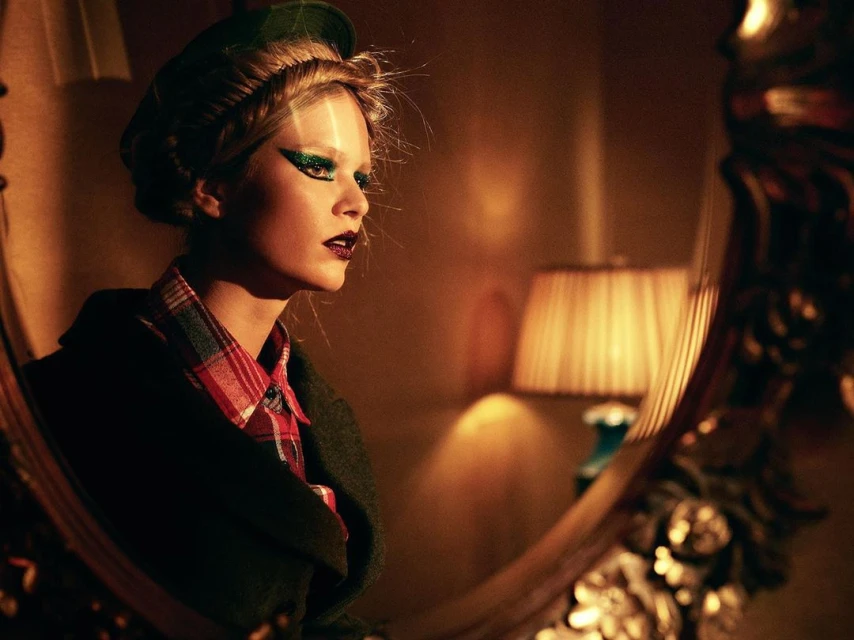 Woman with green glitter eye makeup and dark lipstick gazes near a partially visible mirror, wearing a hat and plaid shirt indoors.