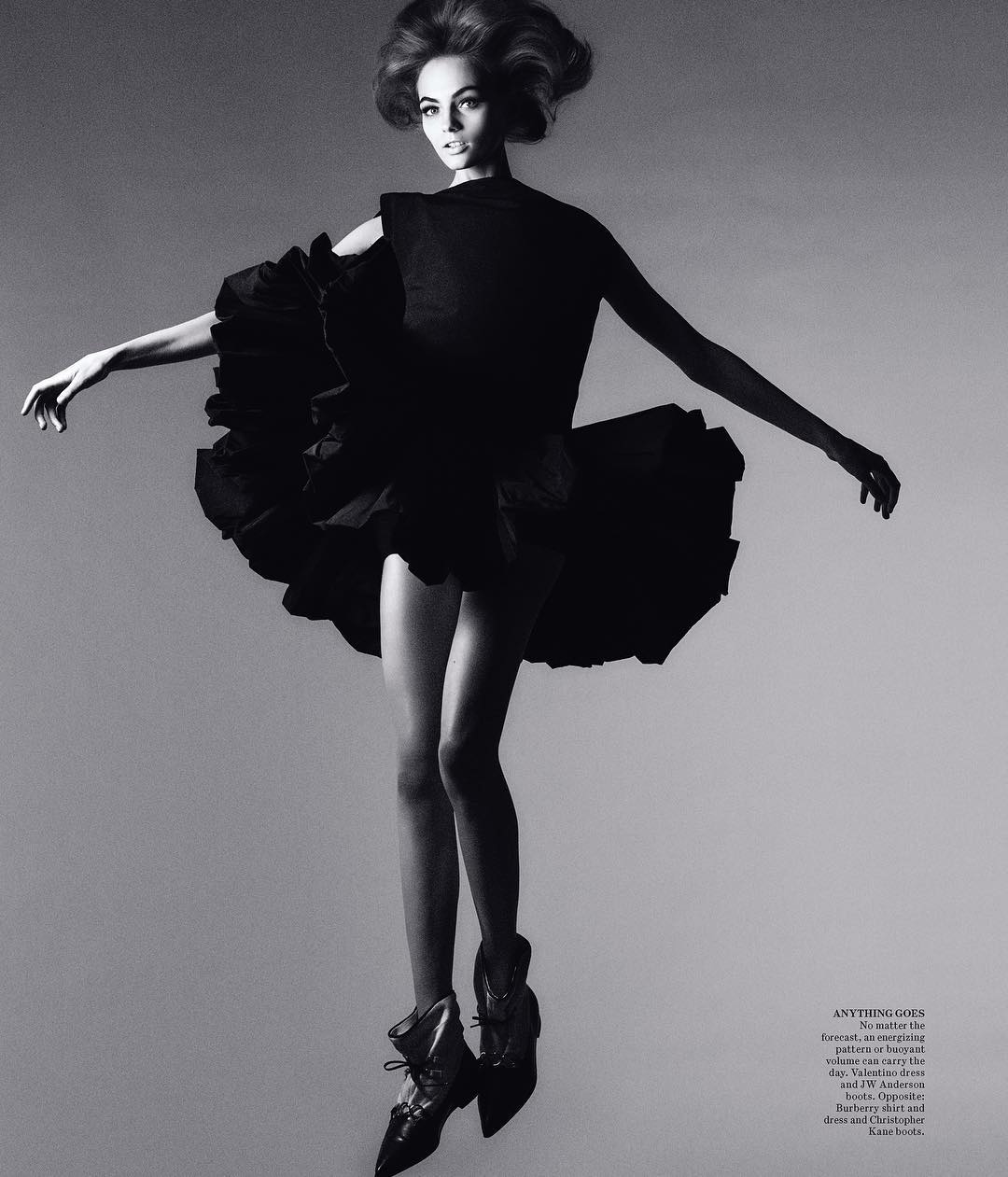 Black and white fashion shot of a woman in a dark dress with ruffles, high heels, and a vintage hairstyle against a gradient grey background.