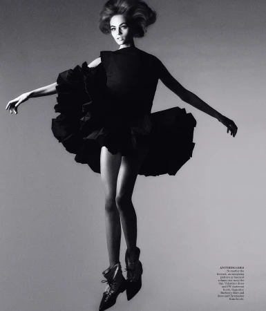 Black and white fashion shot of a woman in a dark dress with ruffles, high heels, and a vintage hairstyle against a gradient grey background.