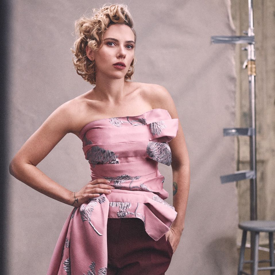 Portrait of a blonde woman in a pink strapless dress with silver botanical pattern posing indoors against a grey backdrop with metal equipment.