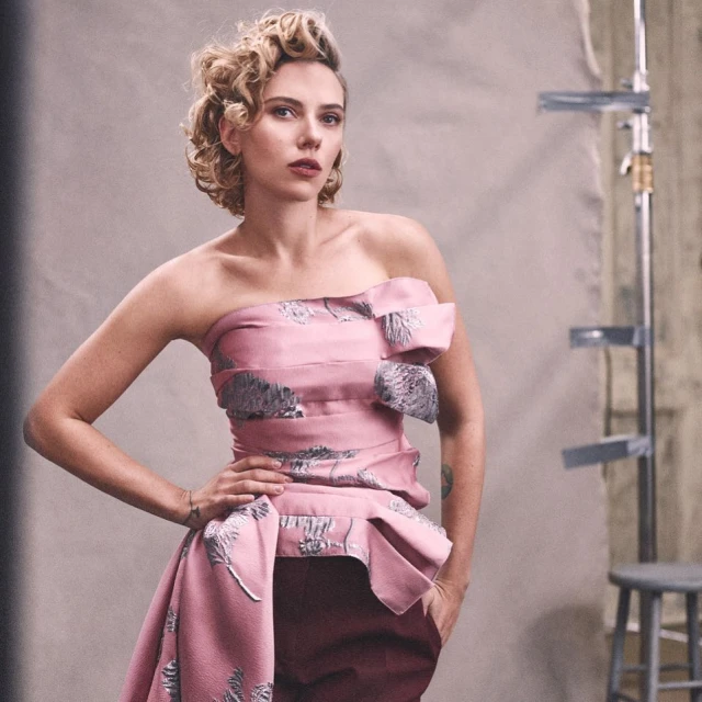 Portrait of a blonde woman in a pink strapless dress with silver botanical pattern posing indoors against a grey backdrop with metal equipment.