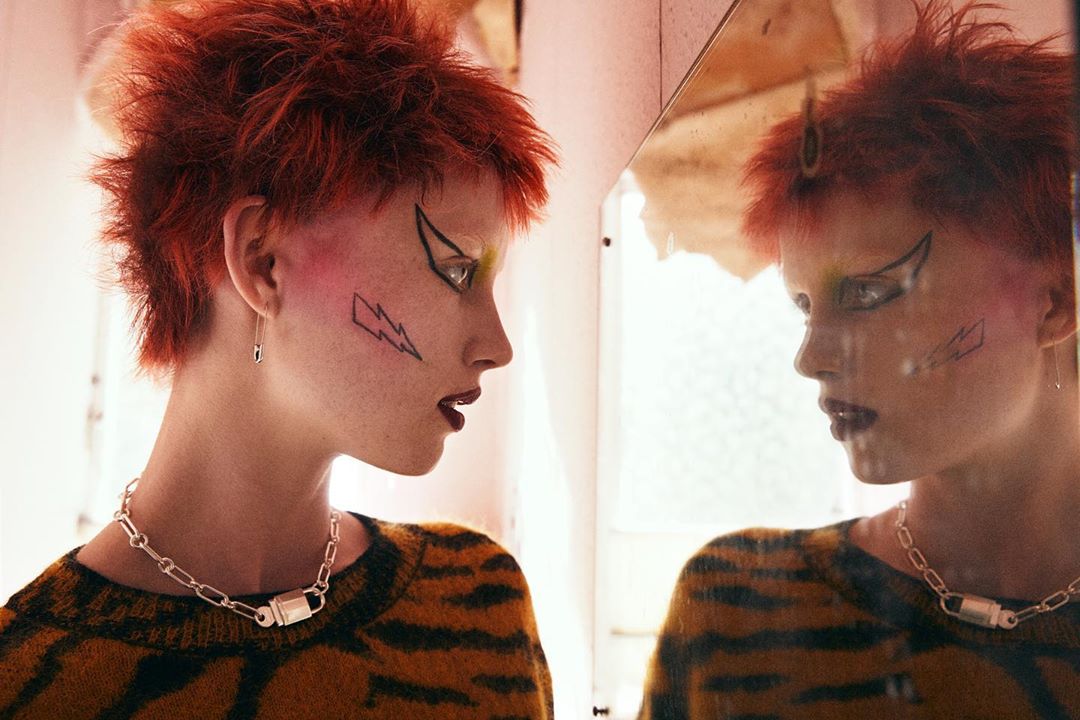 A young adult female with red spiky hair and black graphic eyeliner looking at her reflection wearing a tiger-striped top and chain necklace.