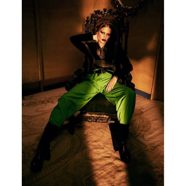 Glamour portrait of a woman with dramatic makeup, seated on an ornate throne, wearing green pants and a black top in a studio setting.