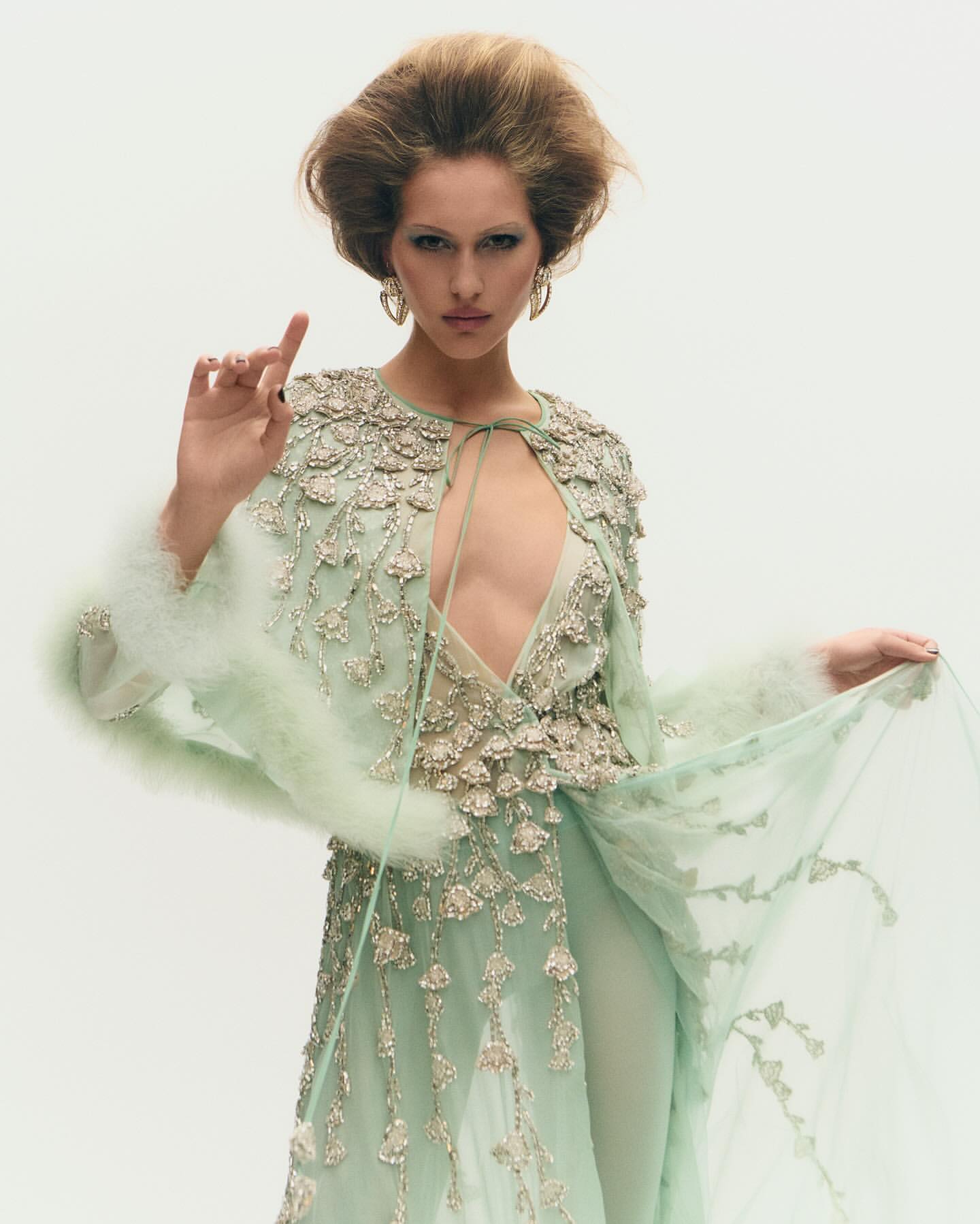 A woman poses in a light green embellished dress with a cape, against a white backdrop, showcasing an editorial fashion aesthetic.