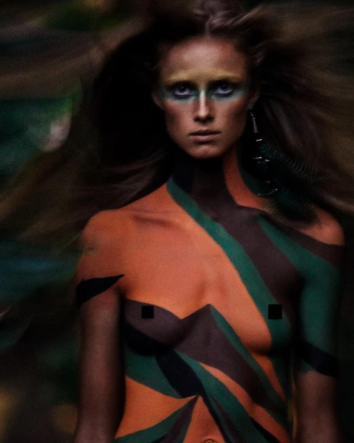 Close-up shot of a person with body paint featuring orange, green, and brown colors, posing in an outdoor environment with blurred foliage.