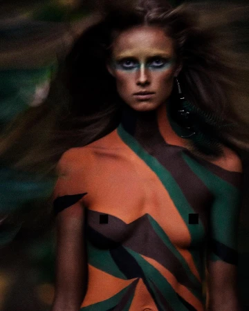 Close-up shot of a person with body paint featuring orange, green, and brown colors, posing in an outdoor environment with blurred foliage.