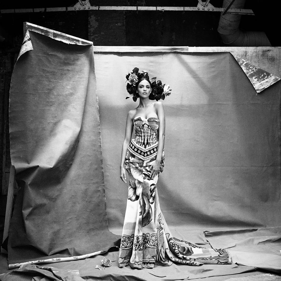 Black and white portrait of a woman in an ornate gown and floral headpiece posing against a textured backdrop in a studio setting.