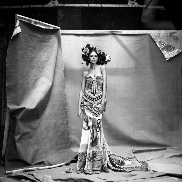 Black and white portrait of a woman in an ornate gown and floral headpiece posing against a textured backdrop in a studio setting.
