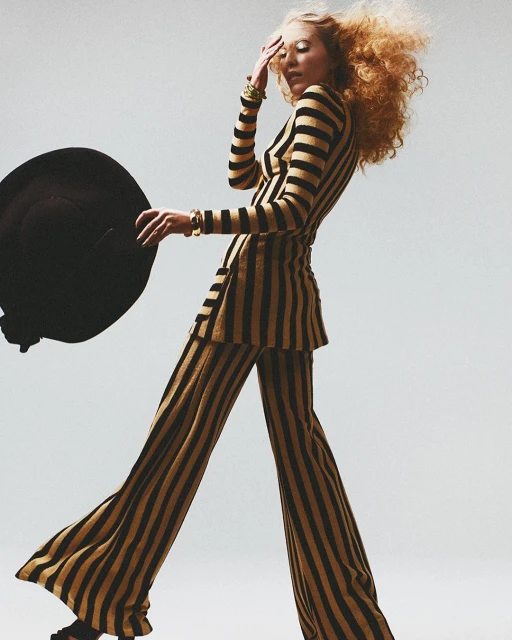 A fashionable adult female posing in a black-and-gold striped suit with wide-leg pants, accented by gold bracelets against a neutral backdrop.